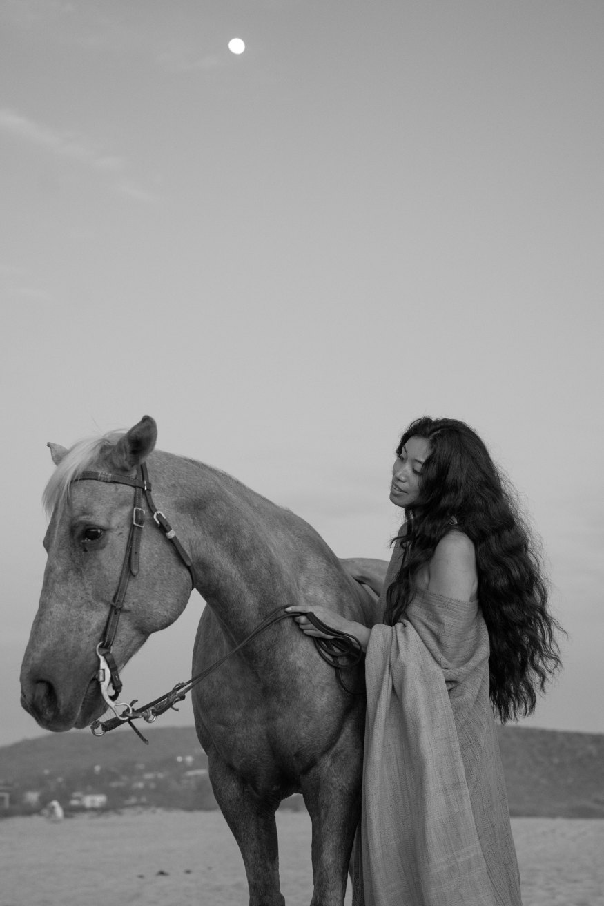 Woman with long dark hair, wrapped in linen, standing beside a grey horse under a full moon